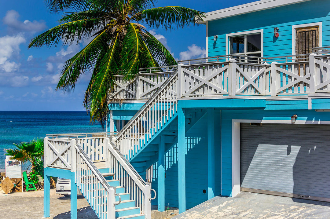 Vibrant blue beach house with palm trees and wooden stairs, overlooking a serene ocean landscape.