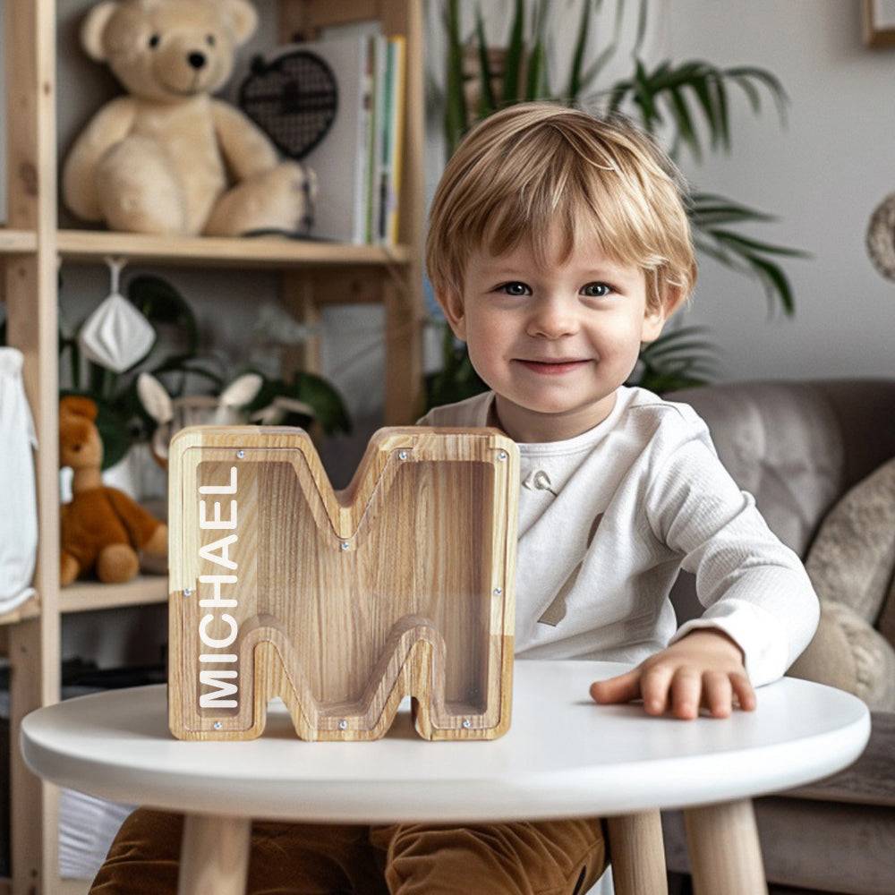 Smiling child with a personalized wooden letter piggy bank shaped like 'M' on a table, showcasing the name Michael.