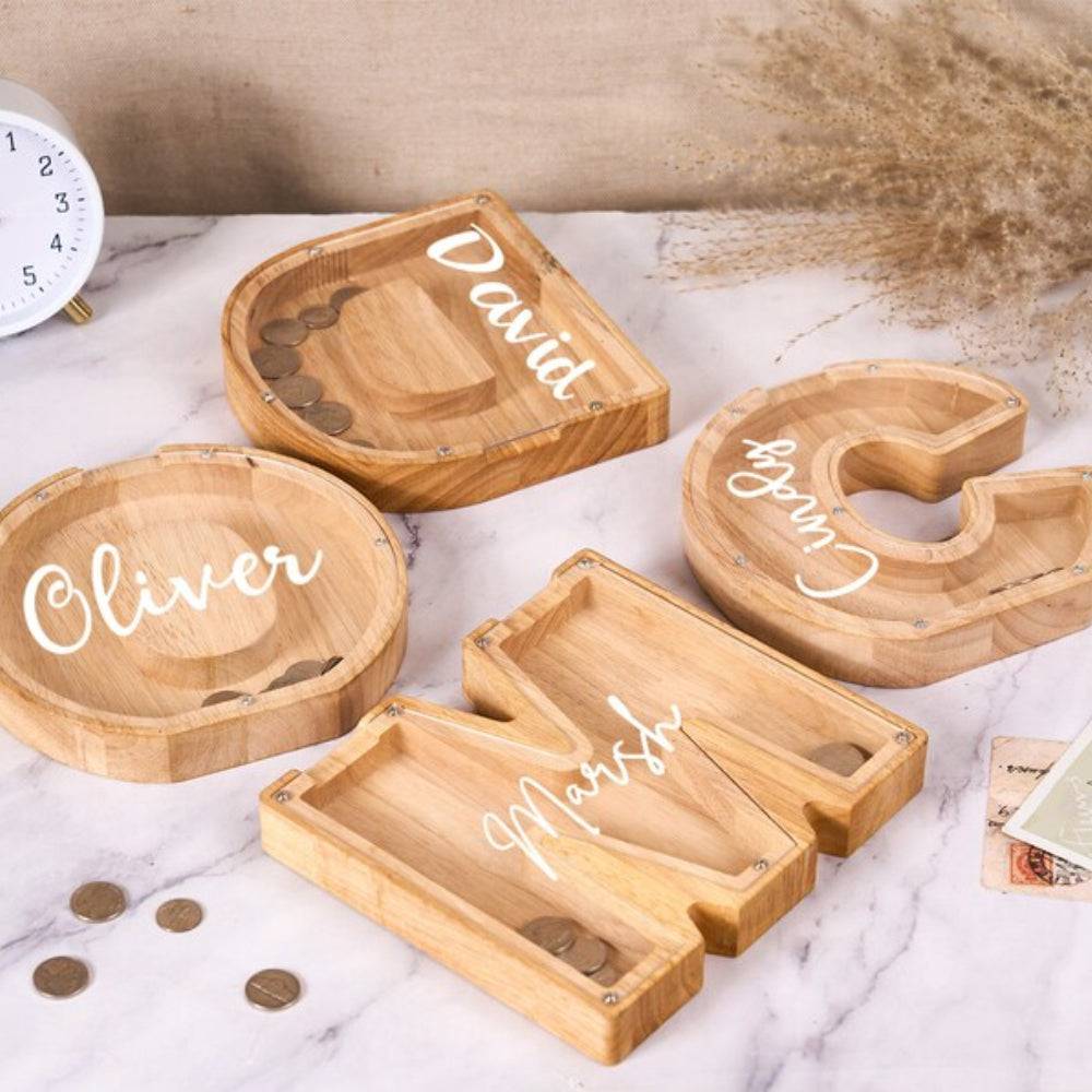 Personalized wooden letter piggy banks with names Oliver, David, and Marsh on a marble surface with coins.